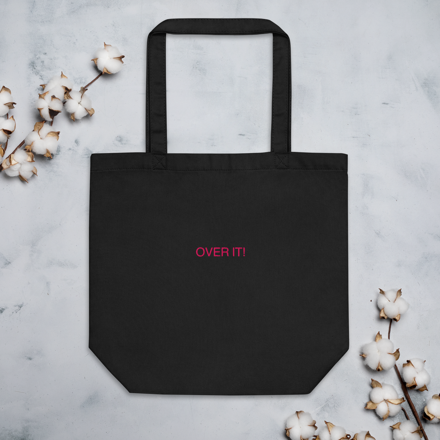 Black Over It slogan embroidered Eco Organic cotton tote bag on a blank surface with cotton plants at the sides.