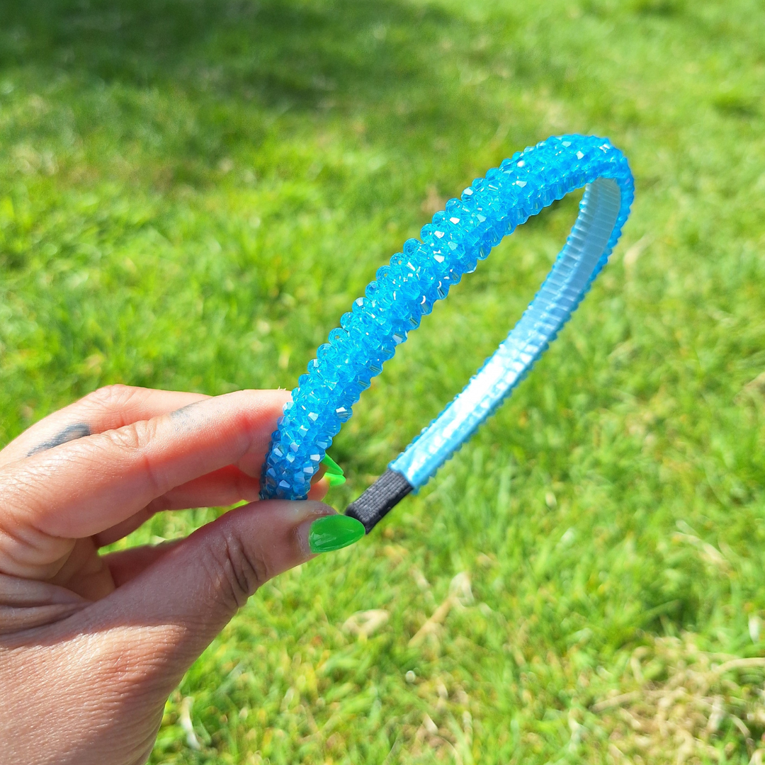 Bright blue rhinestone summer headband being held by a hand in a park in the sun.