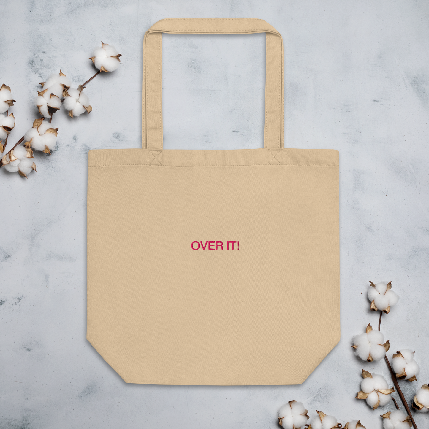 Over It slogan embroidered Eco Organic cotton tote bag on a blank surface with cotton plants around it.