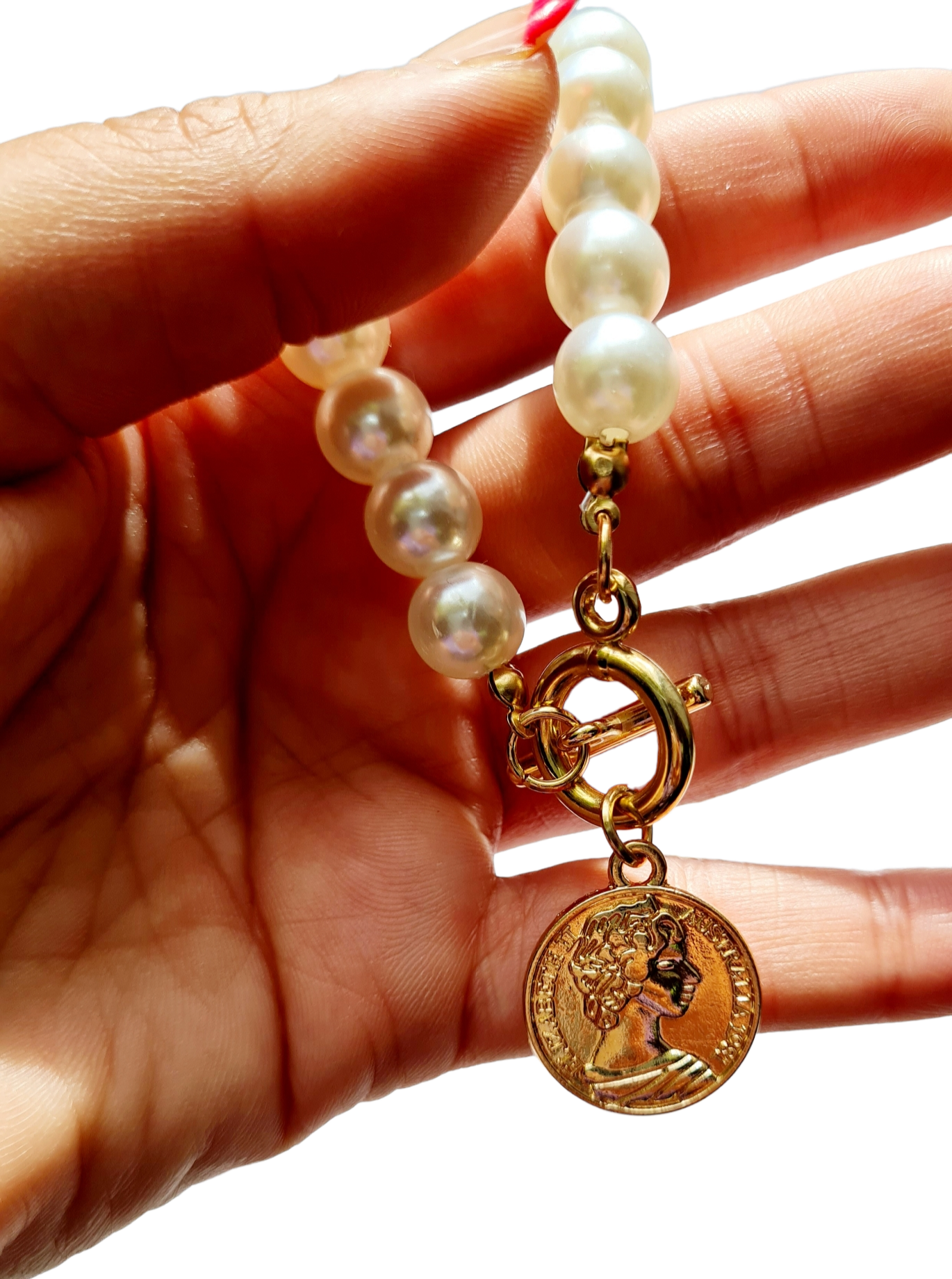 A gold-plated necklace with white pearls and a coin-shaped pendant, displayed on a hand with cool nails on a white background.