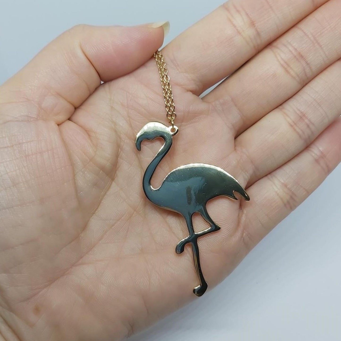Hand holding a gold-plated flamingo charm necklace displayed against a white background.