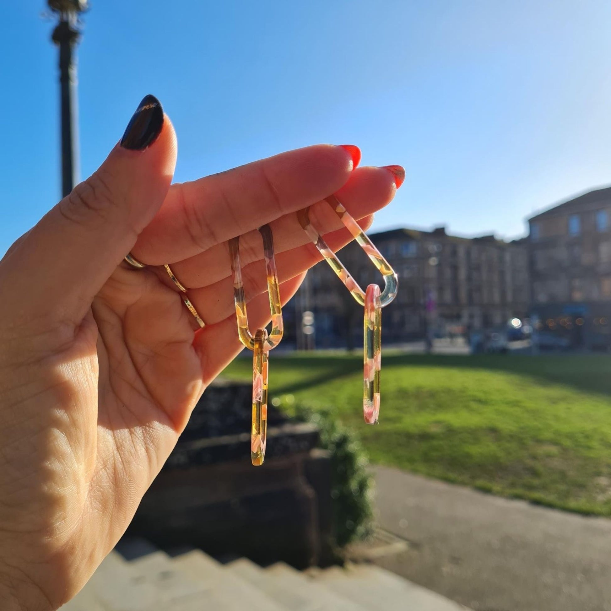 Recycled Summer Rose Link Resin Drop Earrings featuring a geometric rose-shaped design in a translucent yellow and pink resin with gold plating on a hand held up in the sun.