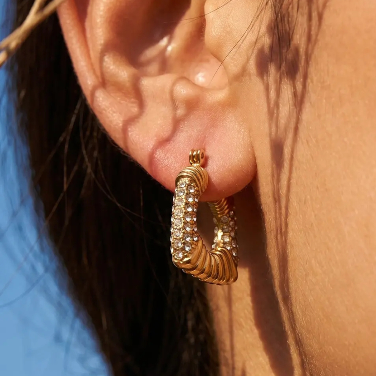 A close-up of a person's ear wearing a gold hoop earring adorned with small clear stones, photographed outdoors in warm sunlight.