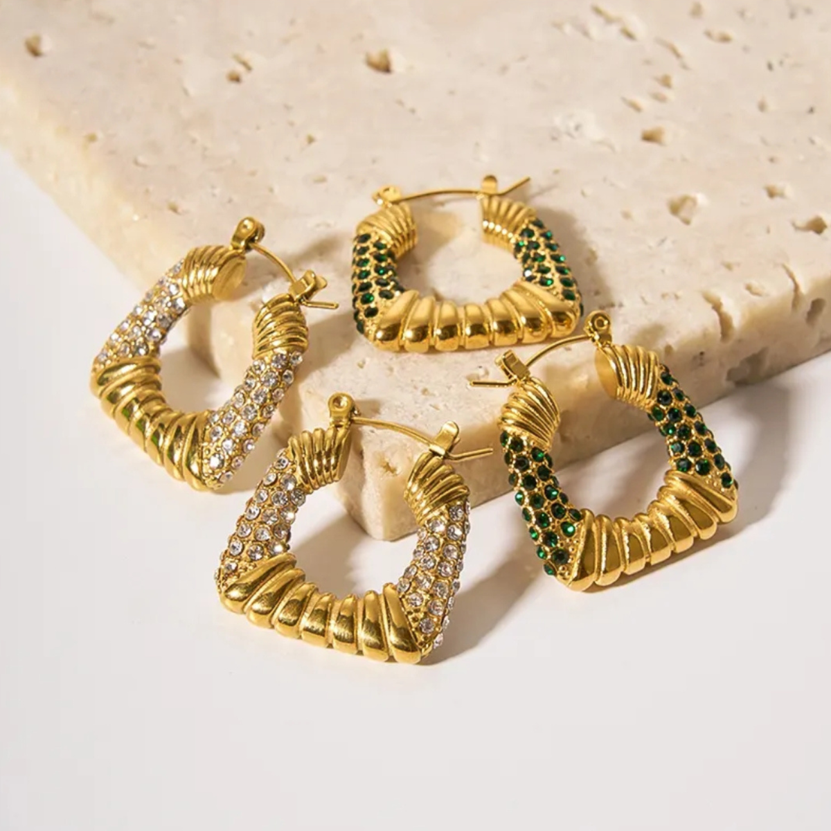 Four gold hoop earrings with twisted rope detailing and either clear pavé stones or green crystal accents, arranged on a light beige stone slab against a white background.