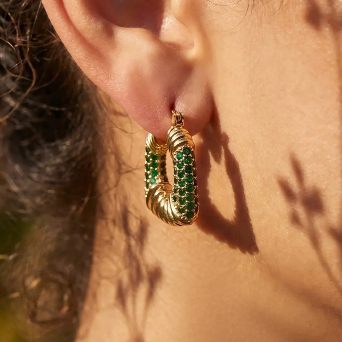 A close-up of a person's ear wearing a gold hoop earring adorned with multiple rows of green gemstones, set against warm sunlight and soft shadows.