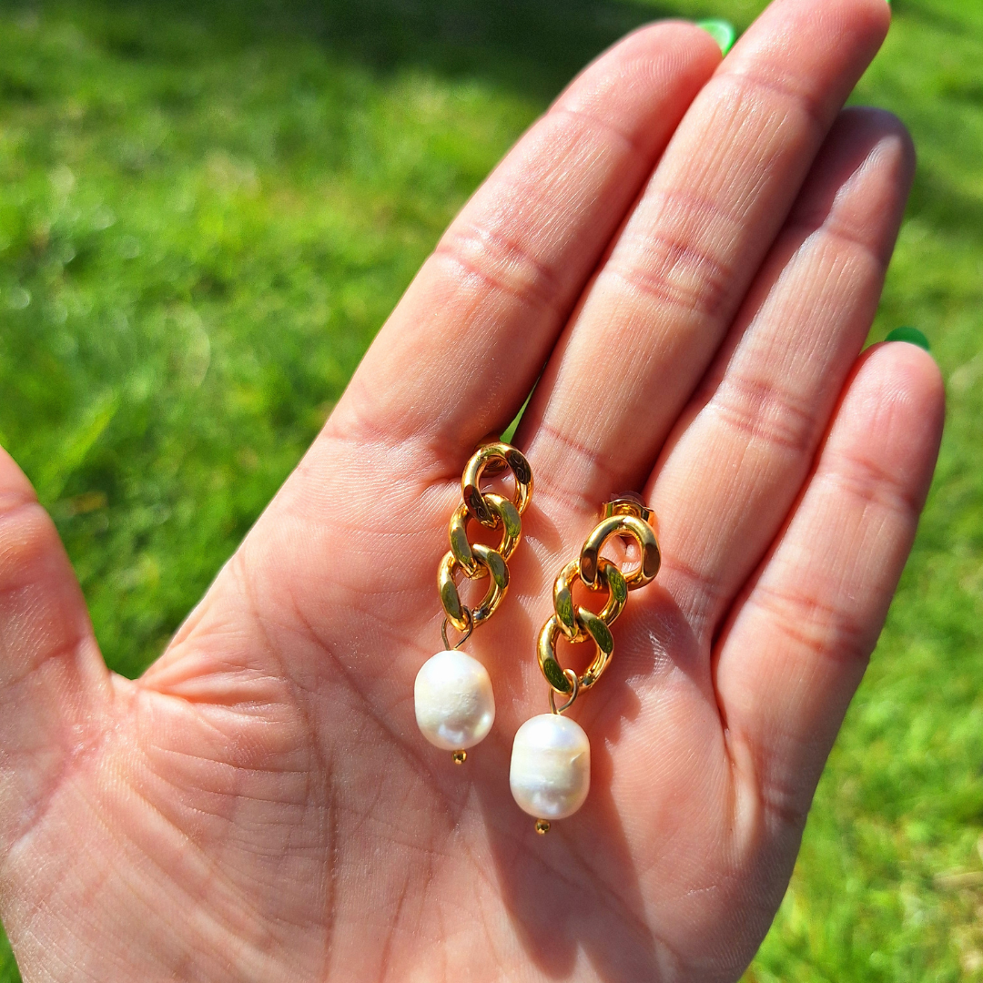 A hand holding a pair of 18K gold plated curb chain earrings with pearl drops, outdoor setting with grass and sunlight visible.