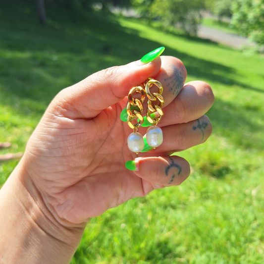 A hand holding a pair of 18K gold plated curb chain earrings with pearl drops, outdoor setting with grass and sunlight visible.