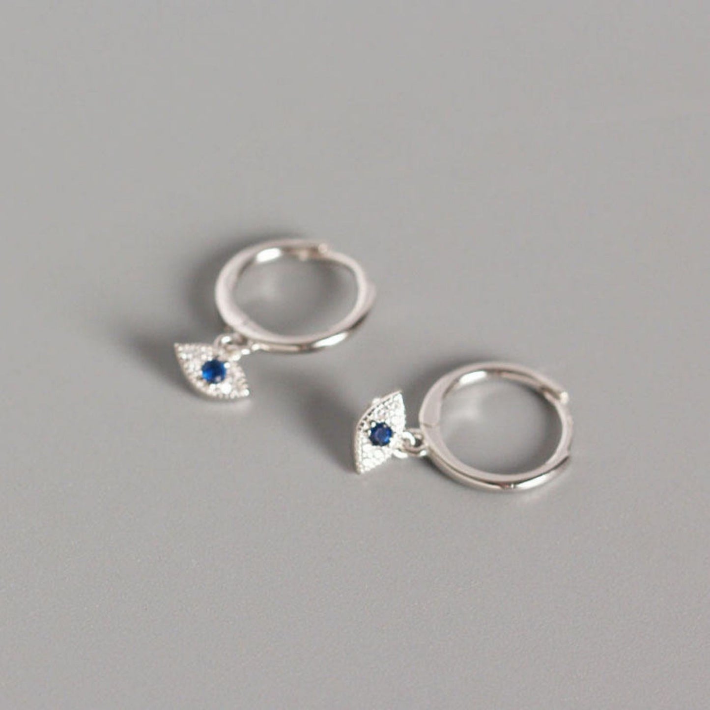 A pair of silver-plated eye-shaped earrings with a sparkling stone in the center, displayed against a grey background.
