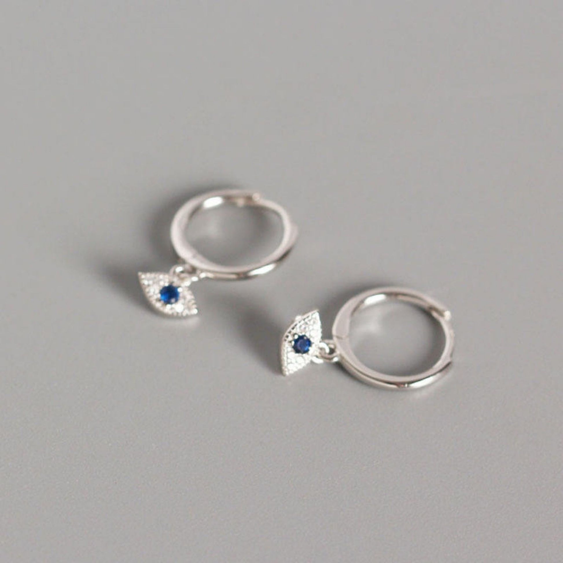 A pair of silver-plated eye-shaped earrings with a sparkling stone in the center, displayed against a grey background.
