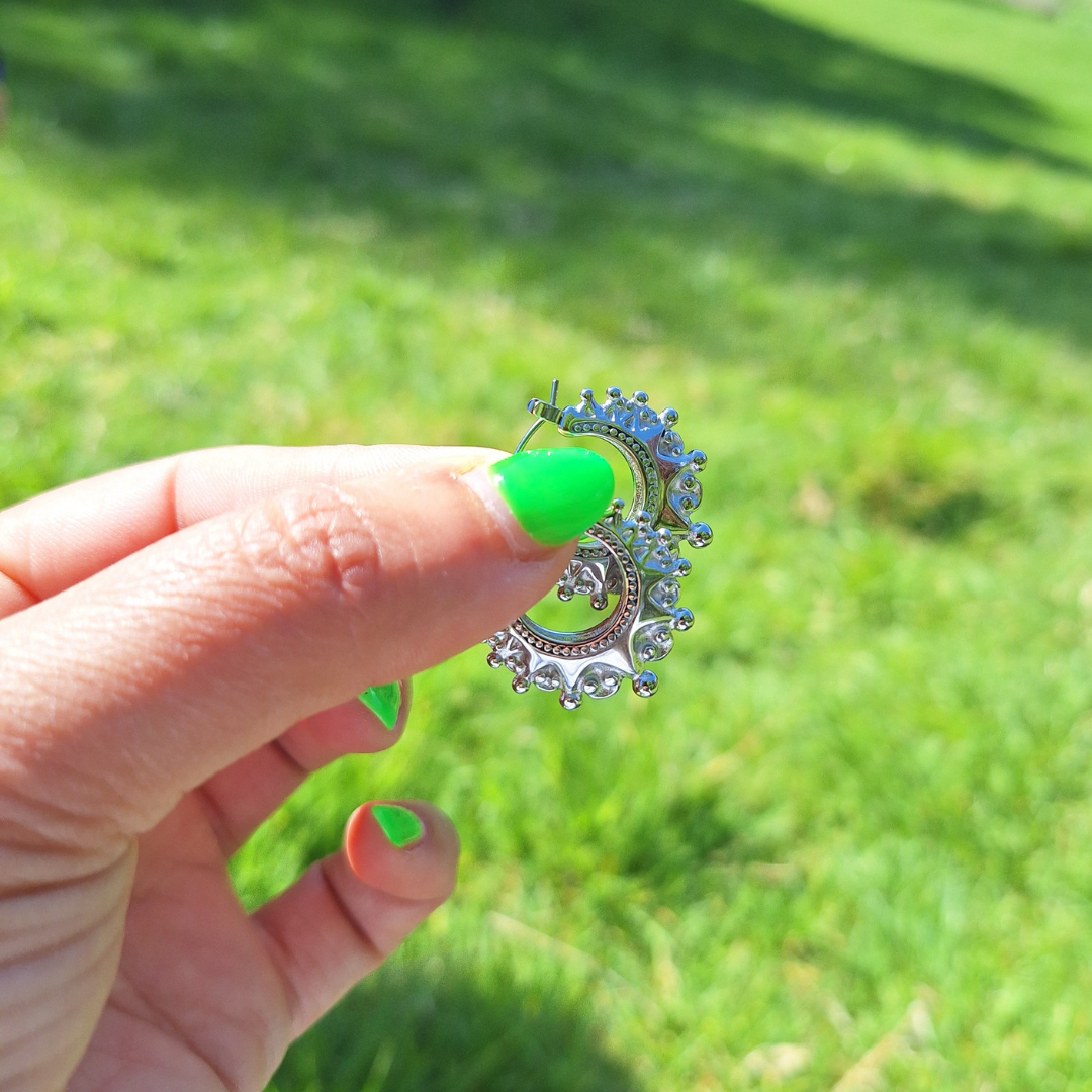A hand with neon green nails holding a pair of silver plaited Arabian creole hoops in the sun.