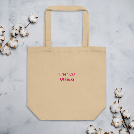 Beige Fresh Out Of Fucks organic cotton Slogan embroidered eco tote bag on a blank surface with cotton plants on the side.