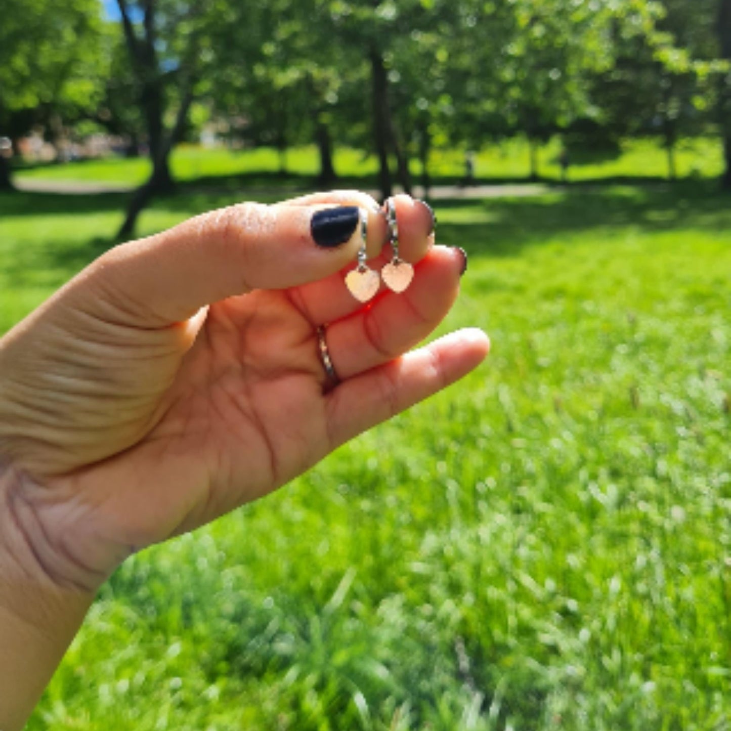 A person holding heart-shaped drop earrings in their hand, with a grassy field in the background.