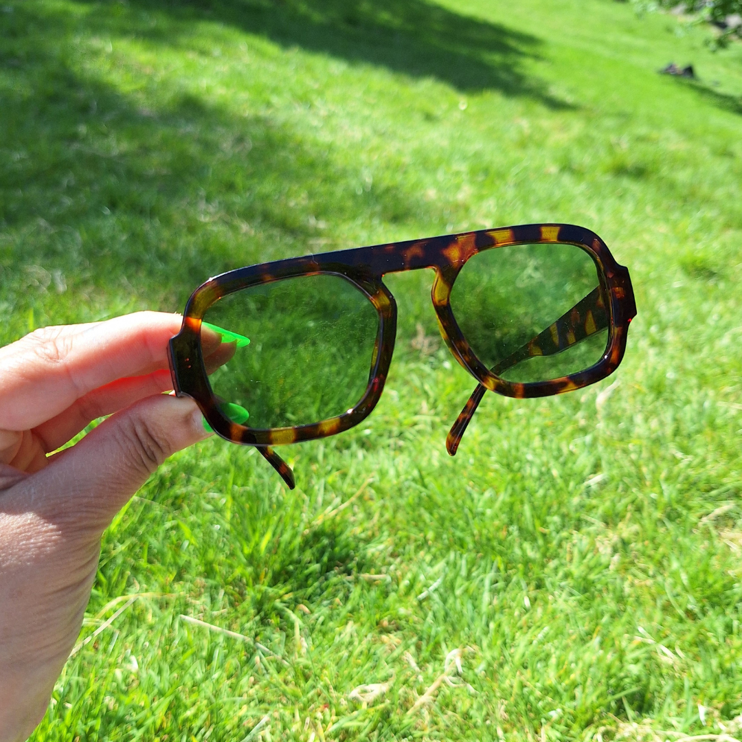Brown Marble Retro Sunnies with Green Lenses being held up in a park in the sun.