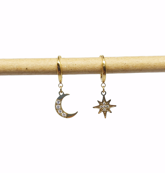 A pair of gold plated celestial earrings, one shaped like a crescent moon and the other like a star, displayed against a wooden backdrop.