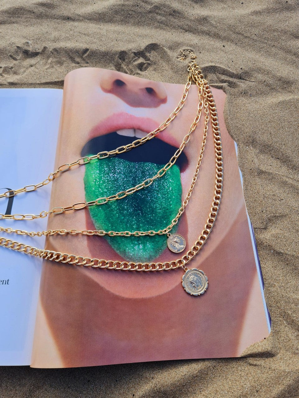 A gold-plated ,multi layer chunky coin necklace displayed on an open magazine on a sandy surface.