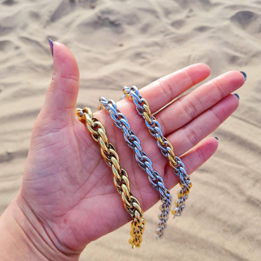 Three chunky twisted bracelets in gold, silver, and mixed metal held in a hand against a sandy background.