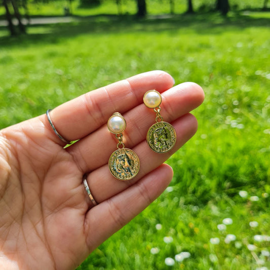 Decadent 14K Gold Plated Coin and Pearl Earrings being held up in the park in the sun.