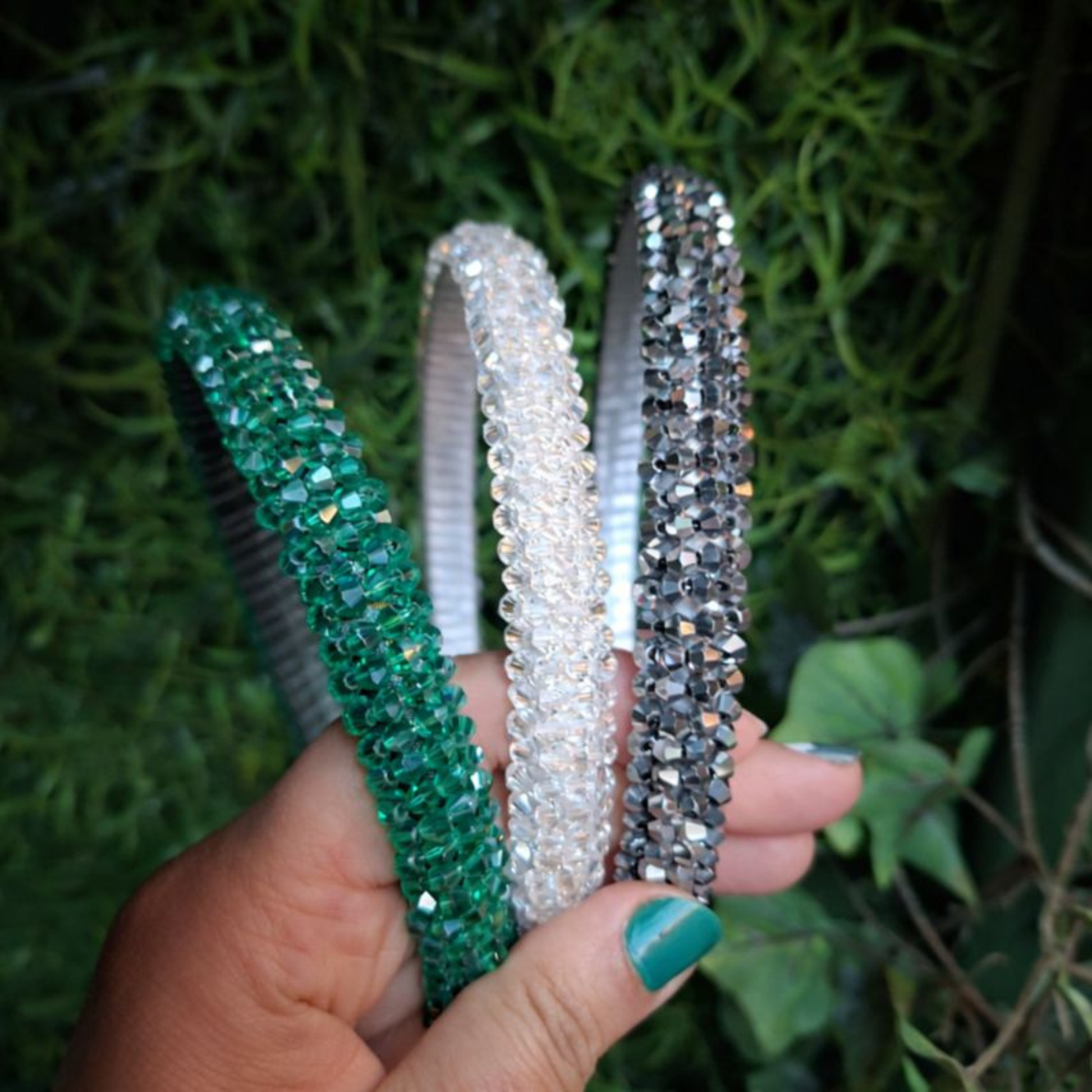 Emerald Green. Clear and Gunmetal grey rhinestone beaded headbands held up in a hand in front of some greenery.
