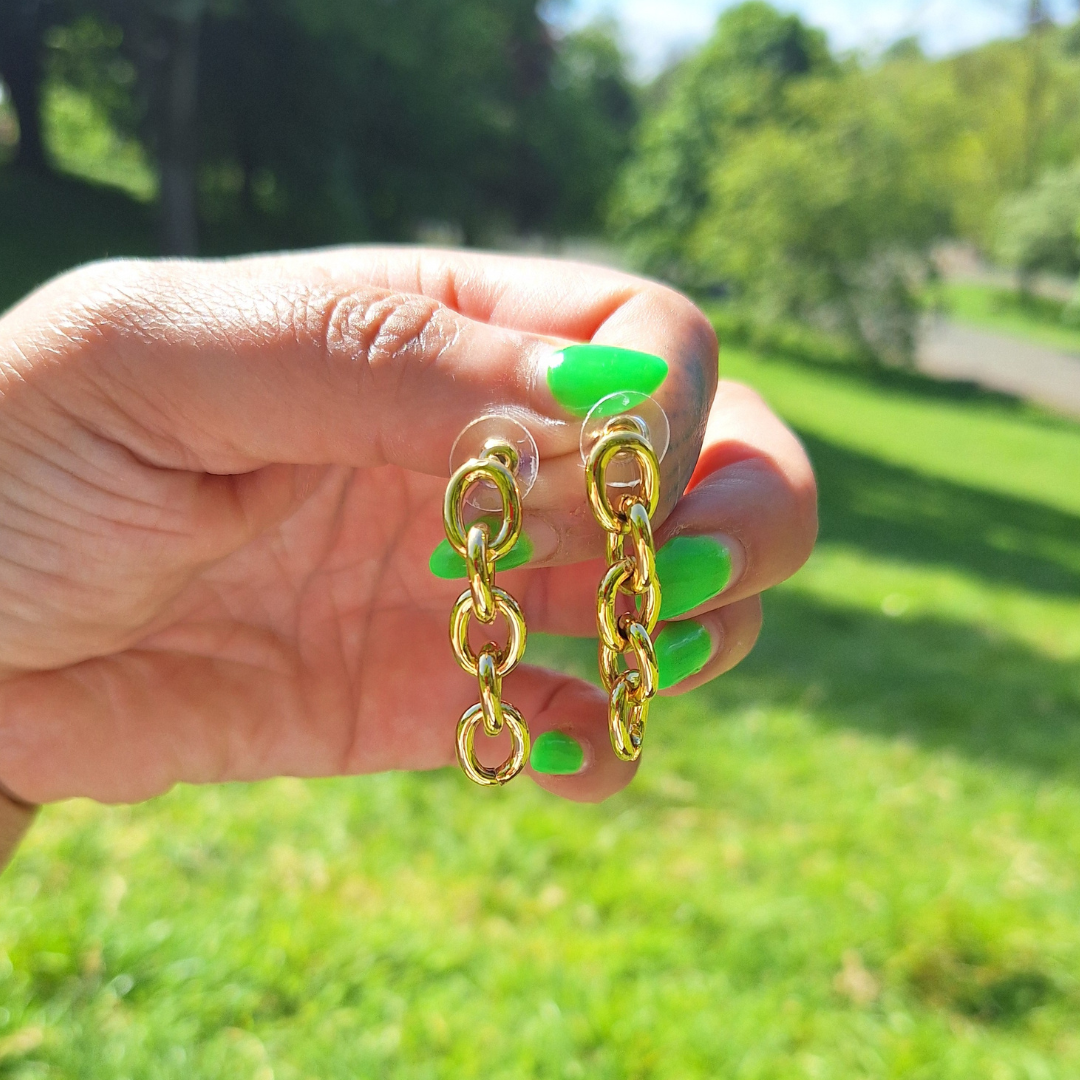A hand holding a gold-plated stainless steel biker chain drop earring outdoors with grass and trees in the background.