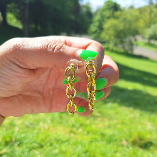 A hand holding a gold-plated stainless steel biker chain drop earring outdoors with grass and trees in the background.