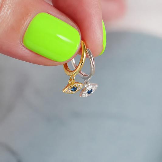 Gold and silver plated huggie earrings with a eye charm with crystal stones and a blue centre stone held by a hand with neon green nail polish.