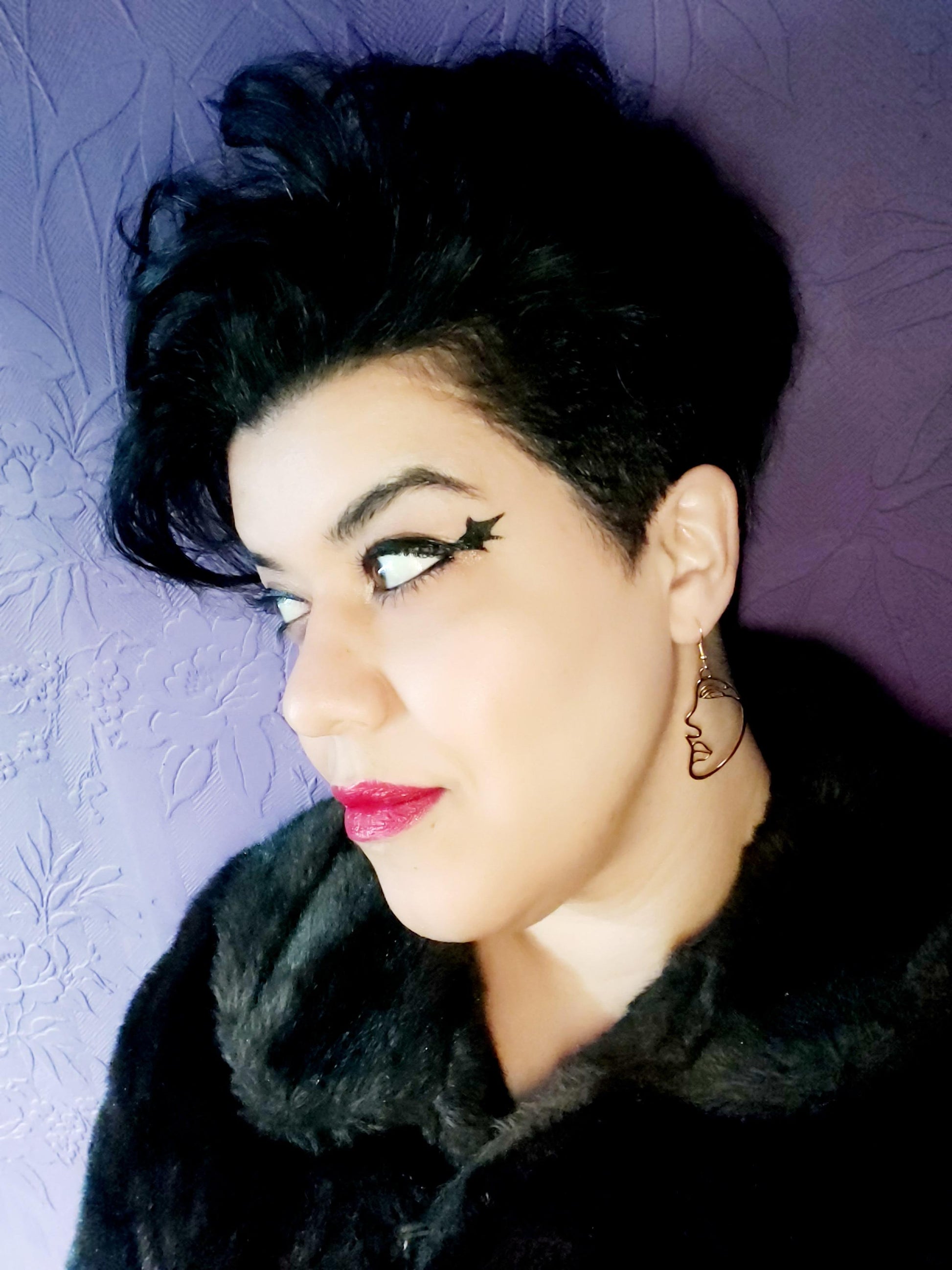 A person wearing gold plated face cut-out earrings, with a purple background.