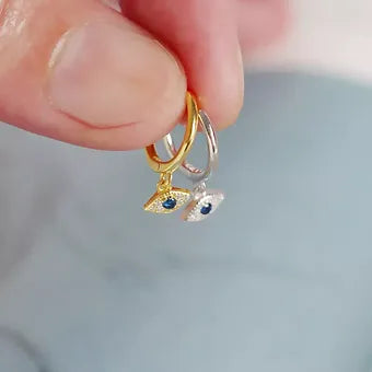 A pair of gold and silver plated evil eye drop hoop earrings with a blue crystal in the eyes held up on a white background.