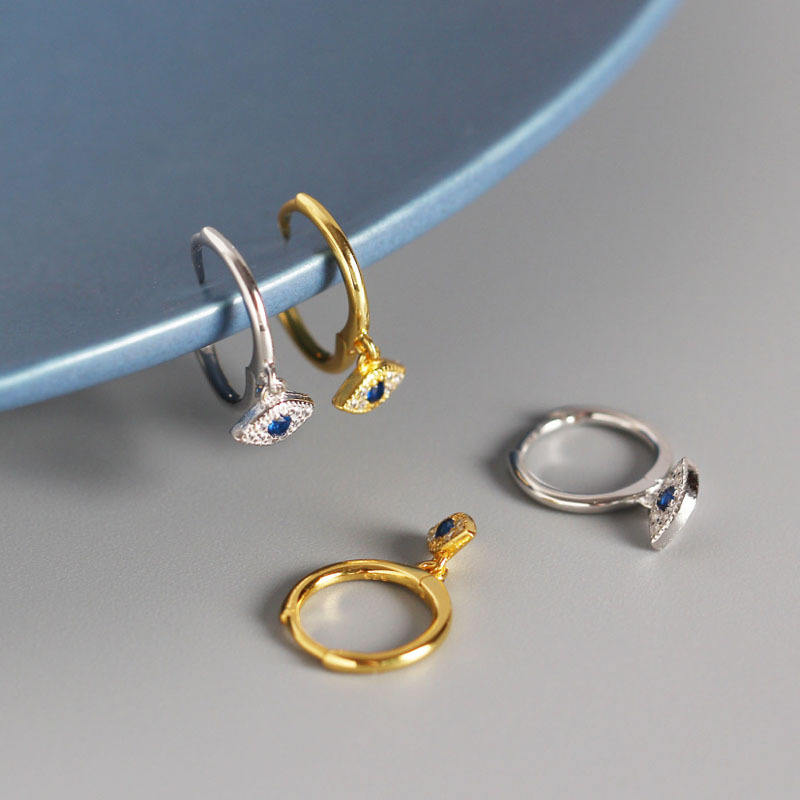 A pair of gold and silver plated evil eye drop hoop earrings with a blue crystal in the eyes on a grey background.