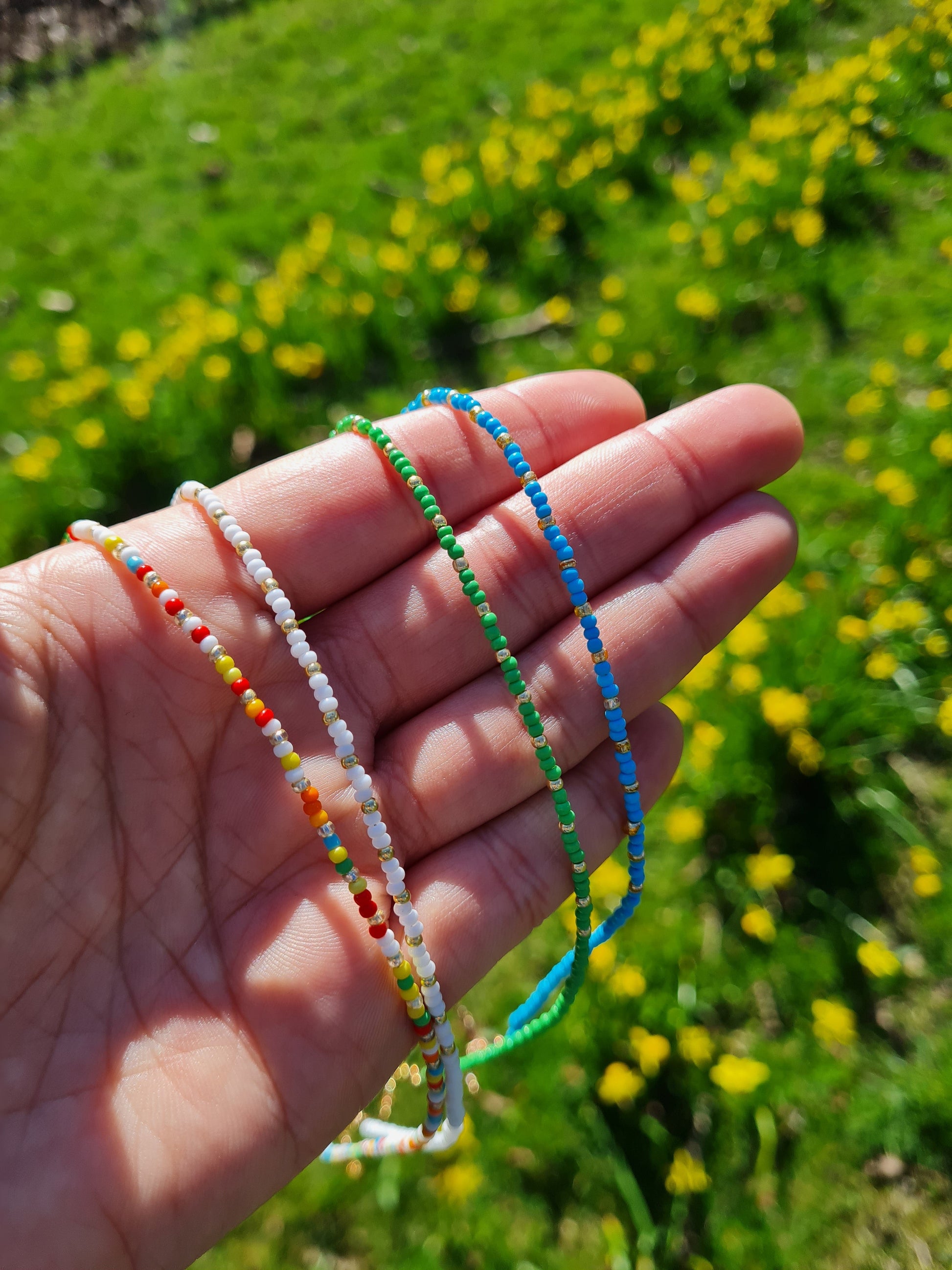 4 multi-coloured beaded necklaces with a gold plated chain and a crystal miyuki bead design, displayed on a hand held up in a park.