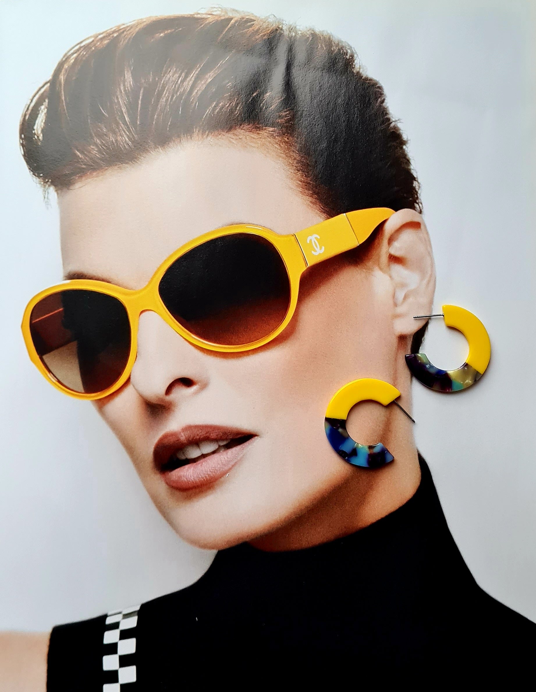 A pair of chunky two-tone mini resin hoop earrings, with one half yellow and the other half multi-colored, laid on a magazine spread of a woman in yellow sunglasses.
