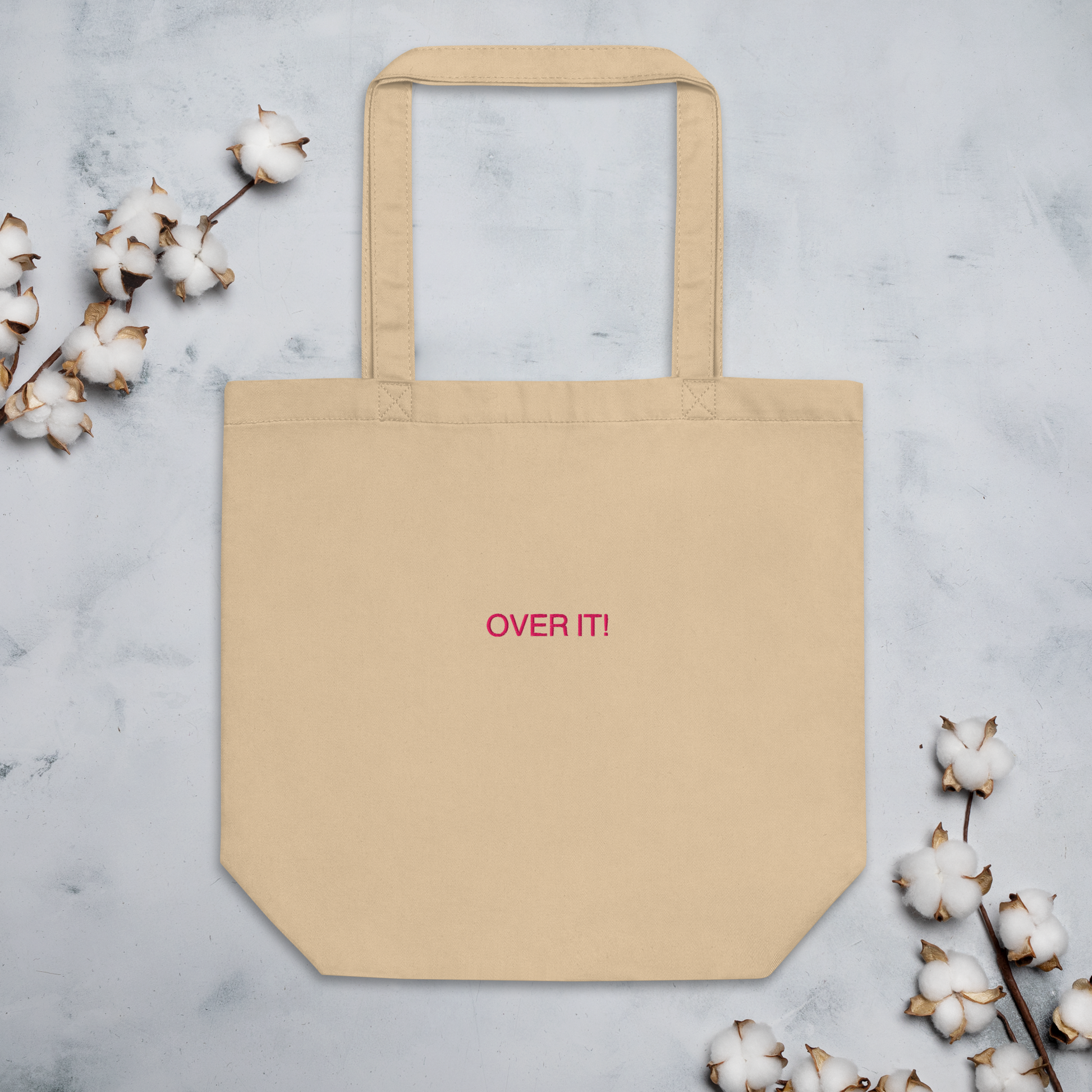 Over It slogan embroidered Eco Organic cotton tote bag on a blank surface with cotton plants around it.