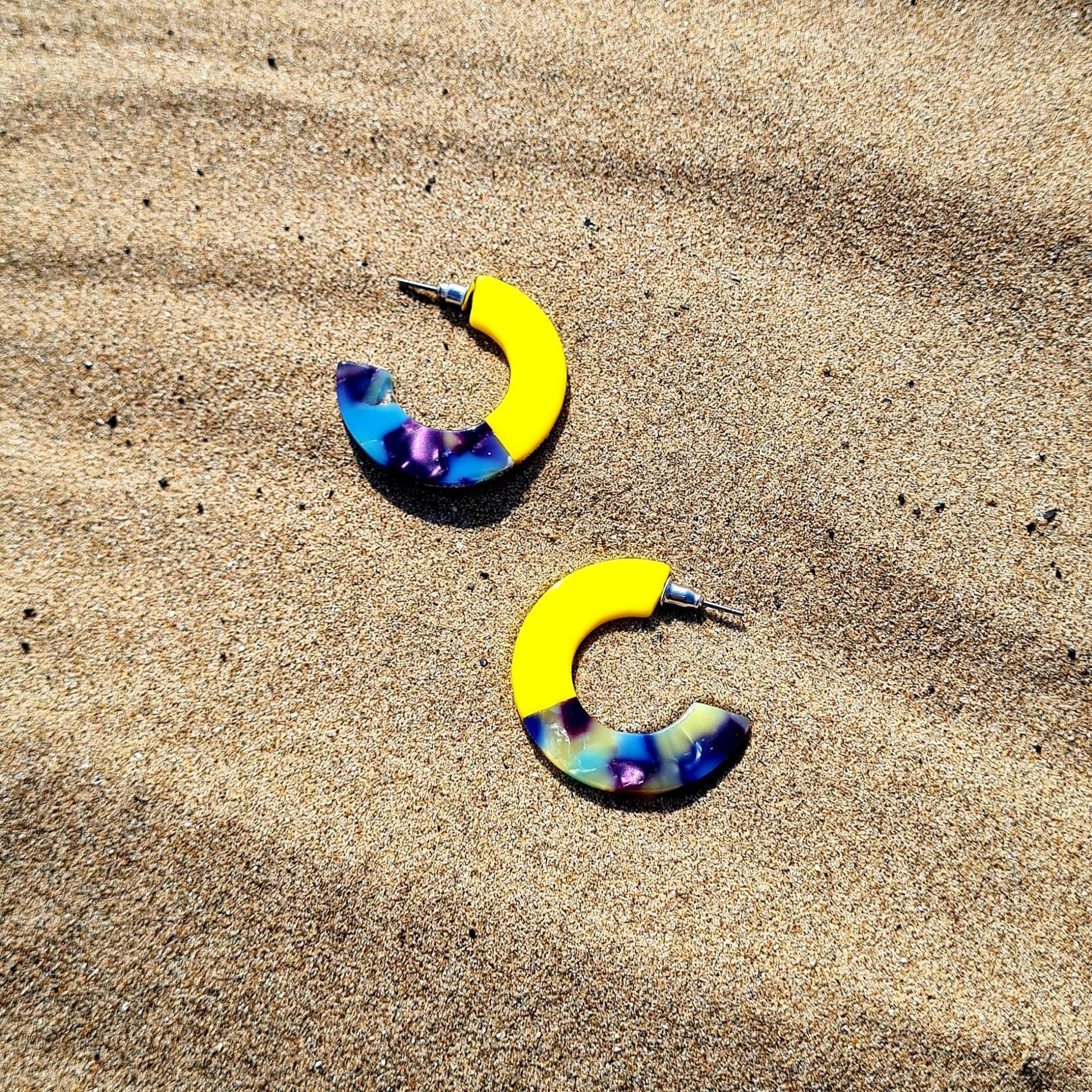 A pair of chunky two-tone mini resin hoop earrings, with one half yellow and the other half multicolored, laid on a sandy surface.