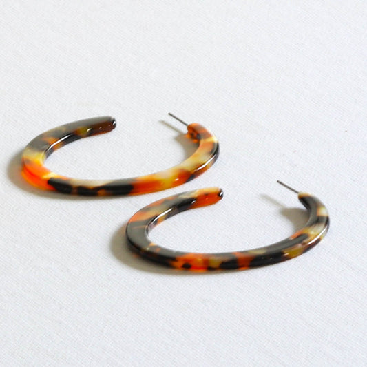 A pair of upcycled flame resin hoop earrings with a gold plated finish, featuring a bold pattern in shades of orange and brown on a white background.