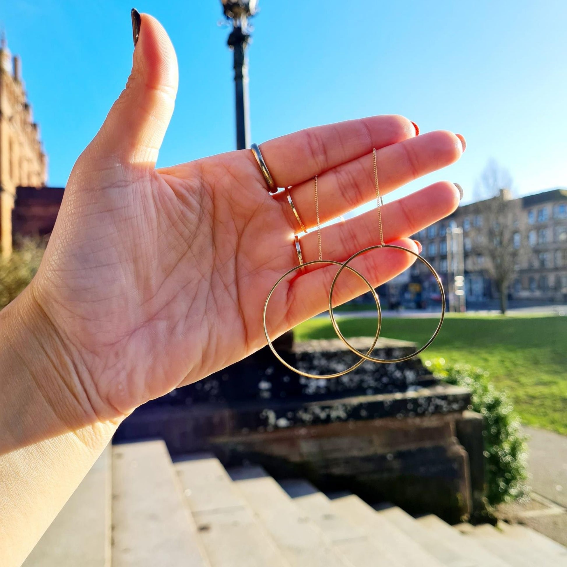 A pair of gold-plated circle pull-through earrings hanging from a hand in the park, showcasing the earrings' full length and circular design.
