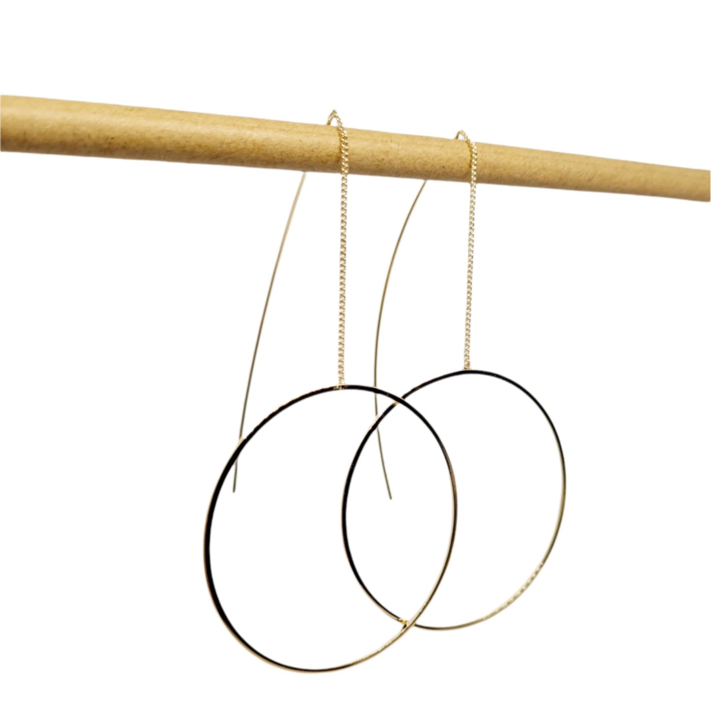 A pair of gold-plated circle pull-through earrings hanging from a wooden display rod, showcasing the earrings' full length and circular design.