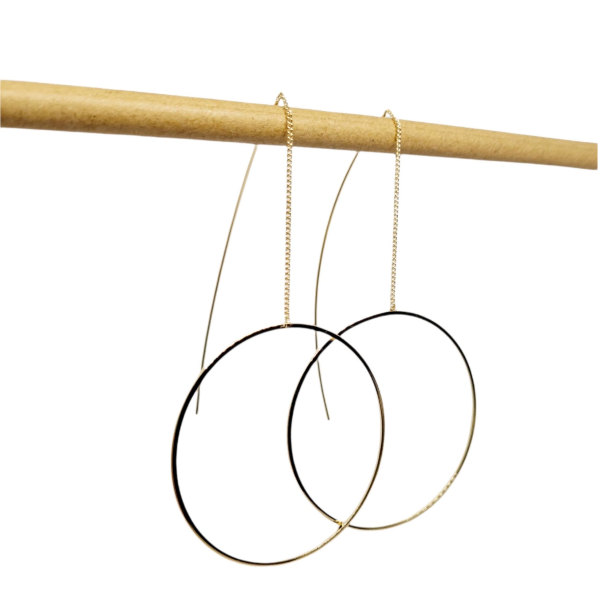 A pair of gold-plated circle pull-through earrings hanging from a wooden display rod, showcasing the earrings' full length and circular design.