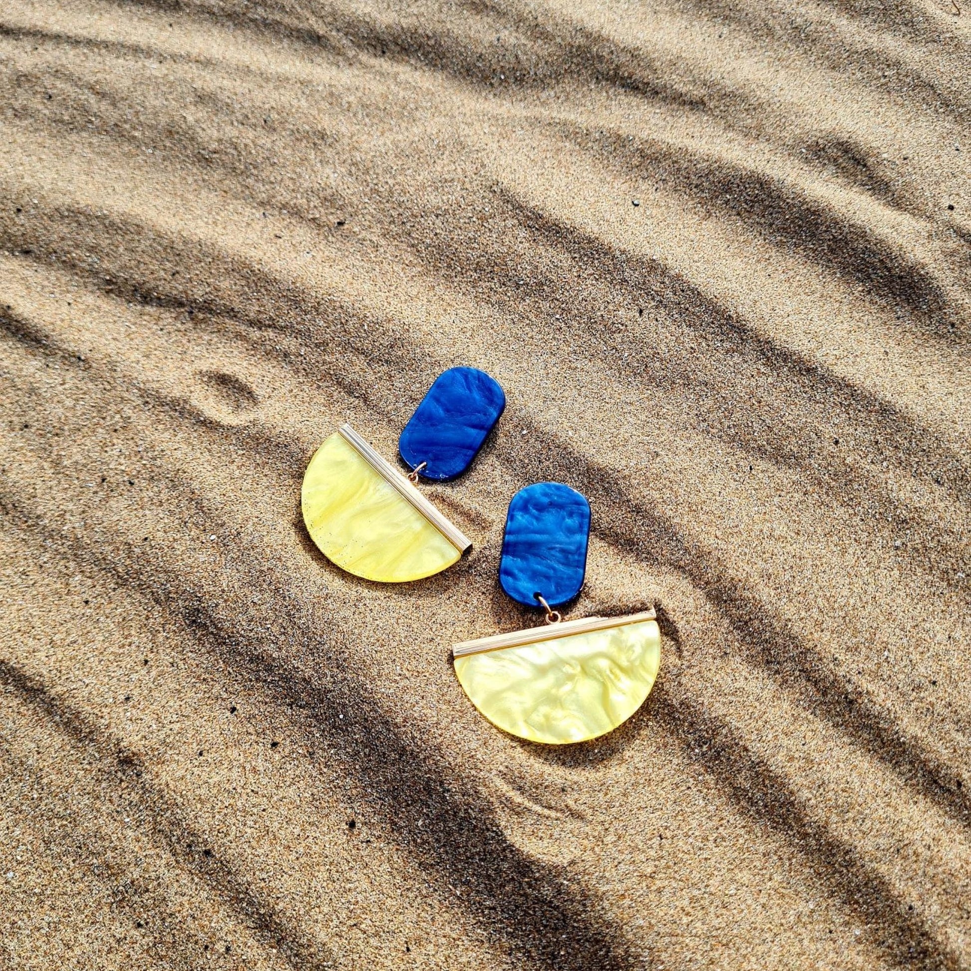 A pair of drop earrings featuring blue and yellow upcycled resin in a geometric design with gold plating on a sandy beach.