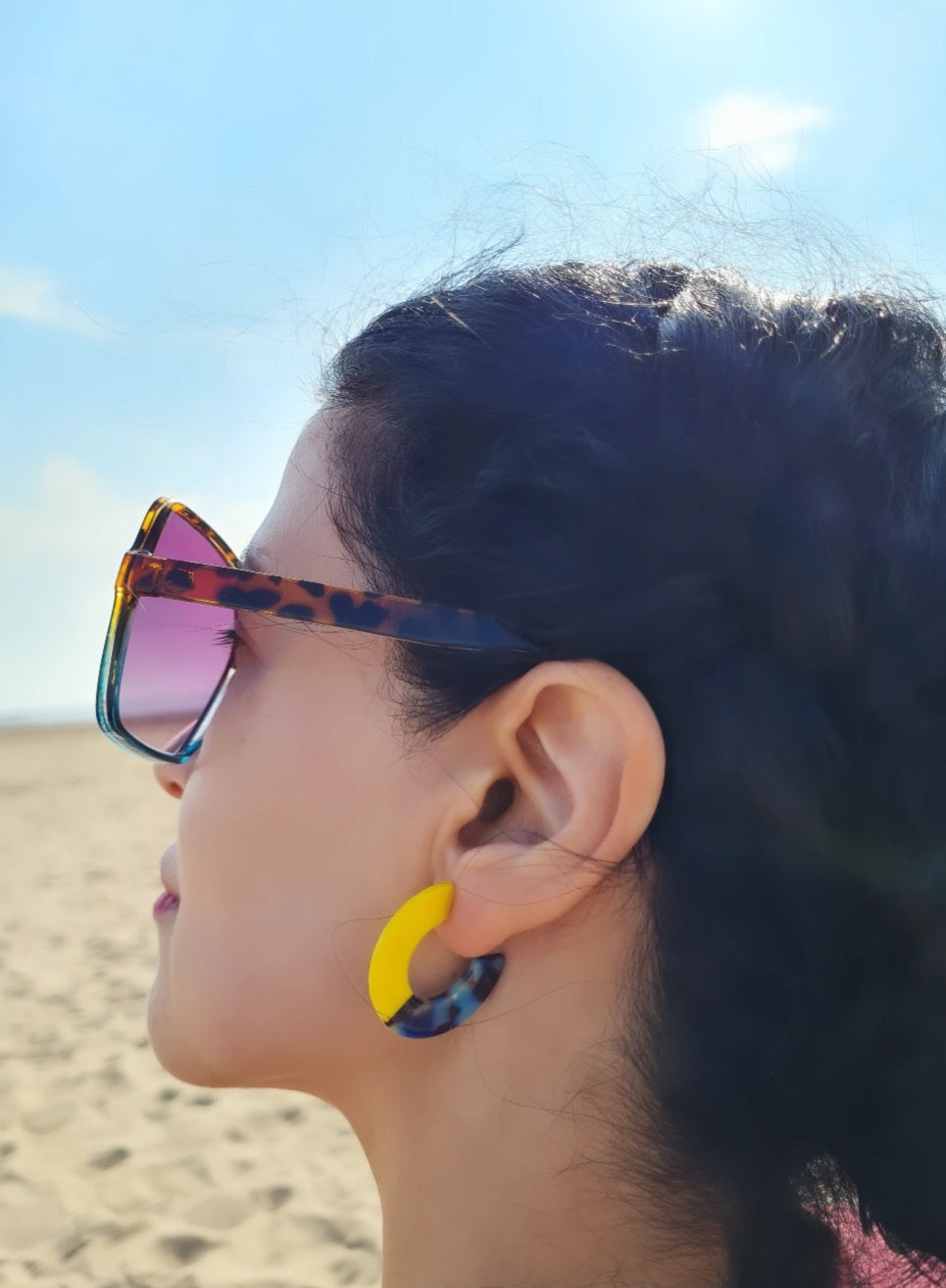 A pair of chunky two-tone mini resin hoop earrings, with one half yellow and the other half multi-colored, on a girl at the beach.