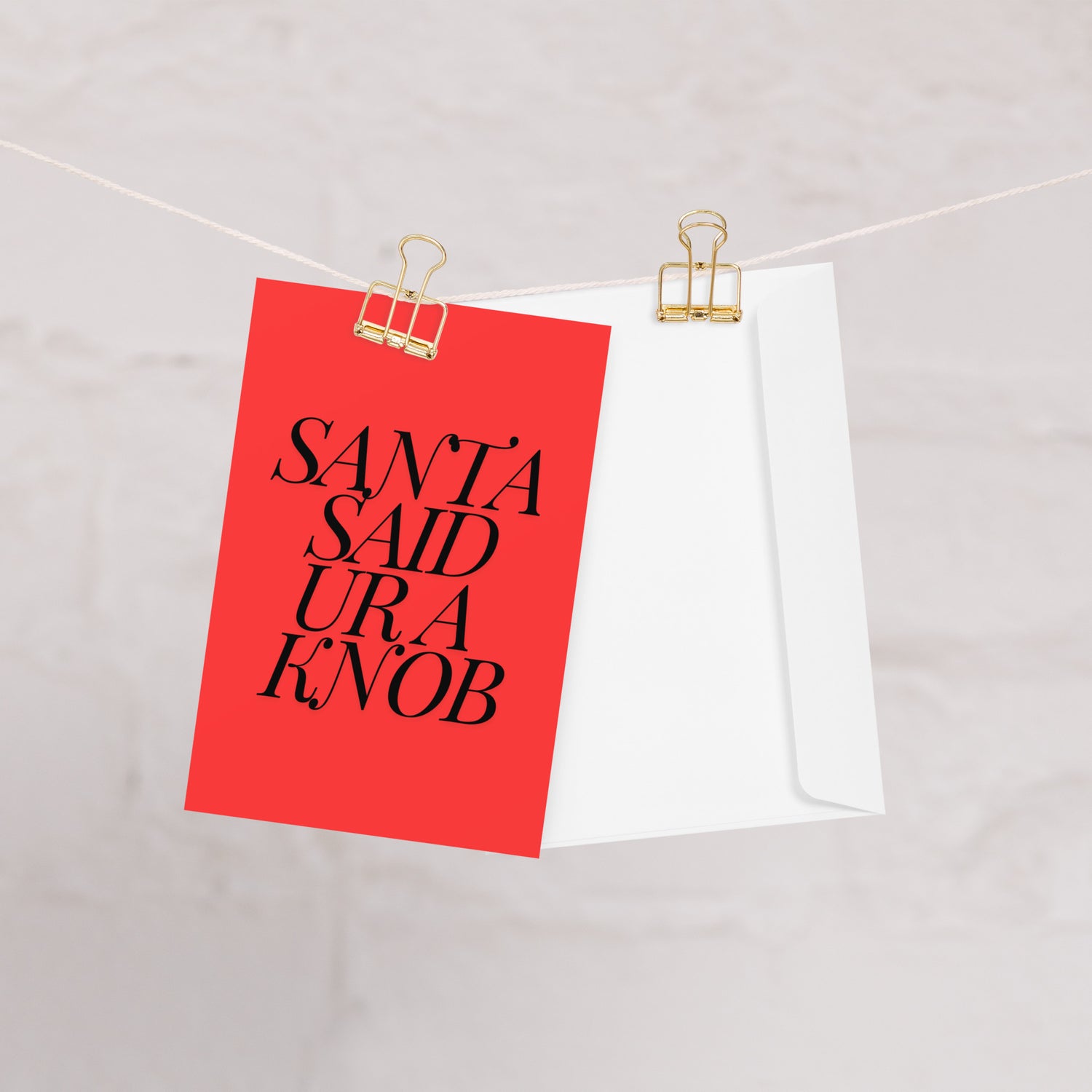 A red card with 'Santa Said Ur a Knob' written in black text, hanging from a gold clip and a complimentary envelope.