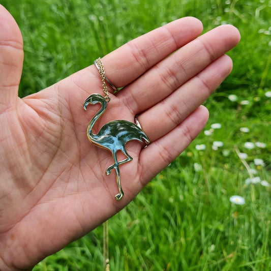Hand holding a gold-plated flamingo charm necklace displayed against a grassy background.