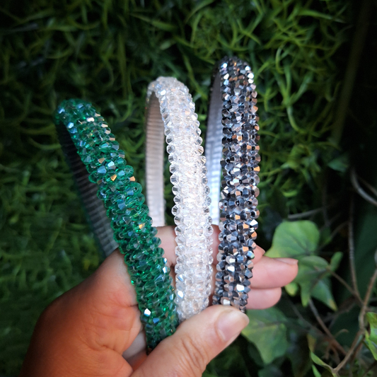 Three decorative headbands in emerald green, white, and charcoal grey held against a green leafy background.