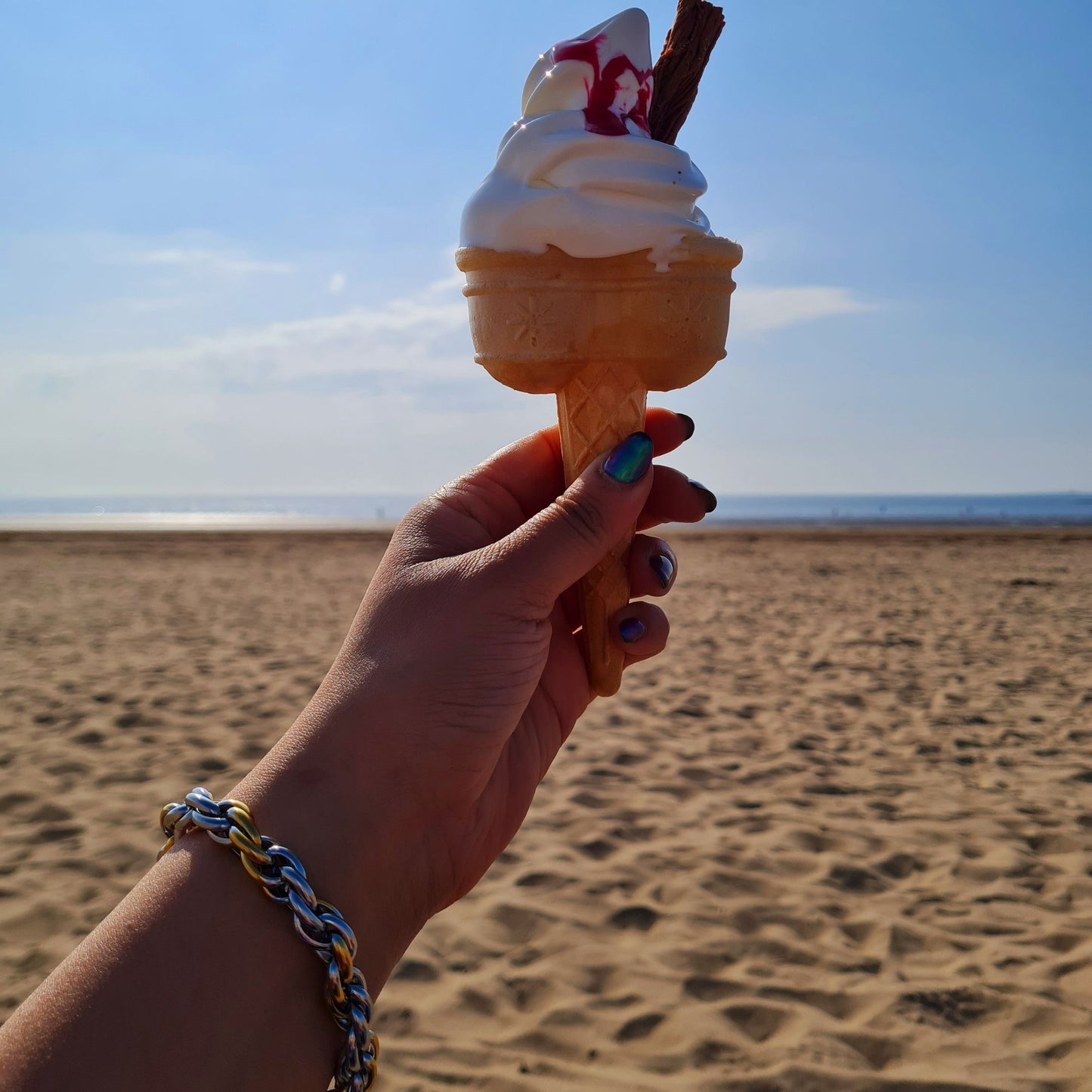 A chunky silver and gold plated twisted bracelet on a wrist at a beach holding an ice-cream cone.
