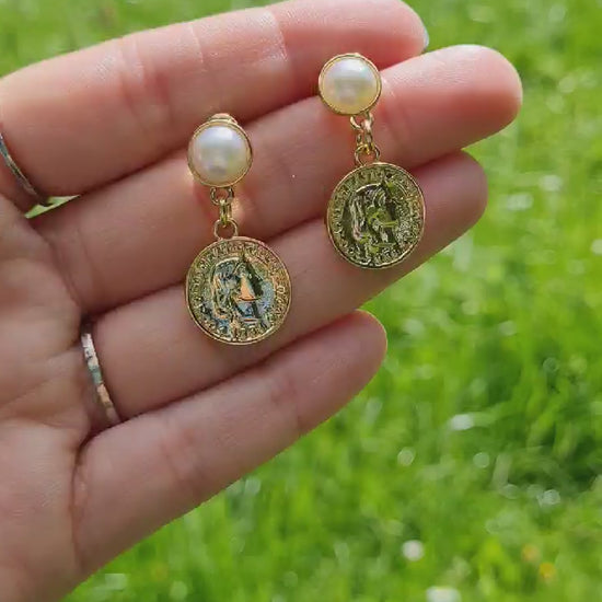 Decadent 14K Gold Plated Coin and Pearl Earrings being held up in the park in the sun.
