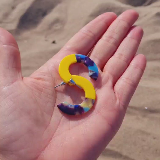 A pair of chunky two-tone mini resin hoop earrings, with one half yellow and the other half multi-colored, laid on a hand on a beach.