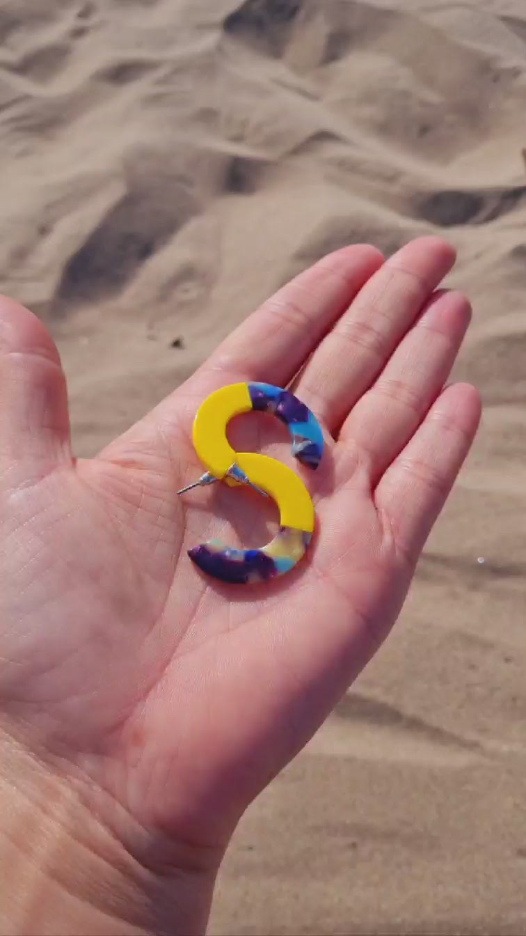 A pair of chunky two-tone mini resin hoop earrings, with one half yellow and the other half multi-colored, laid on a hand on a beach.