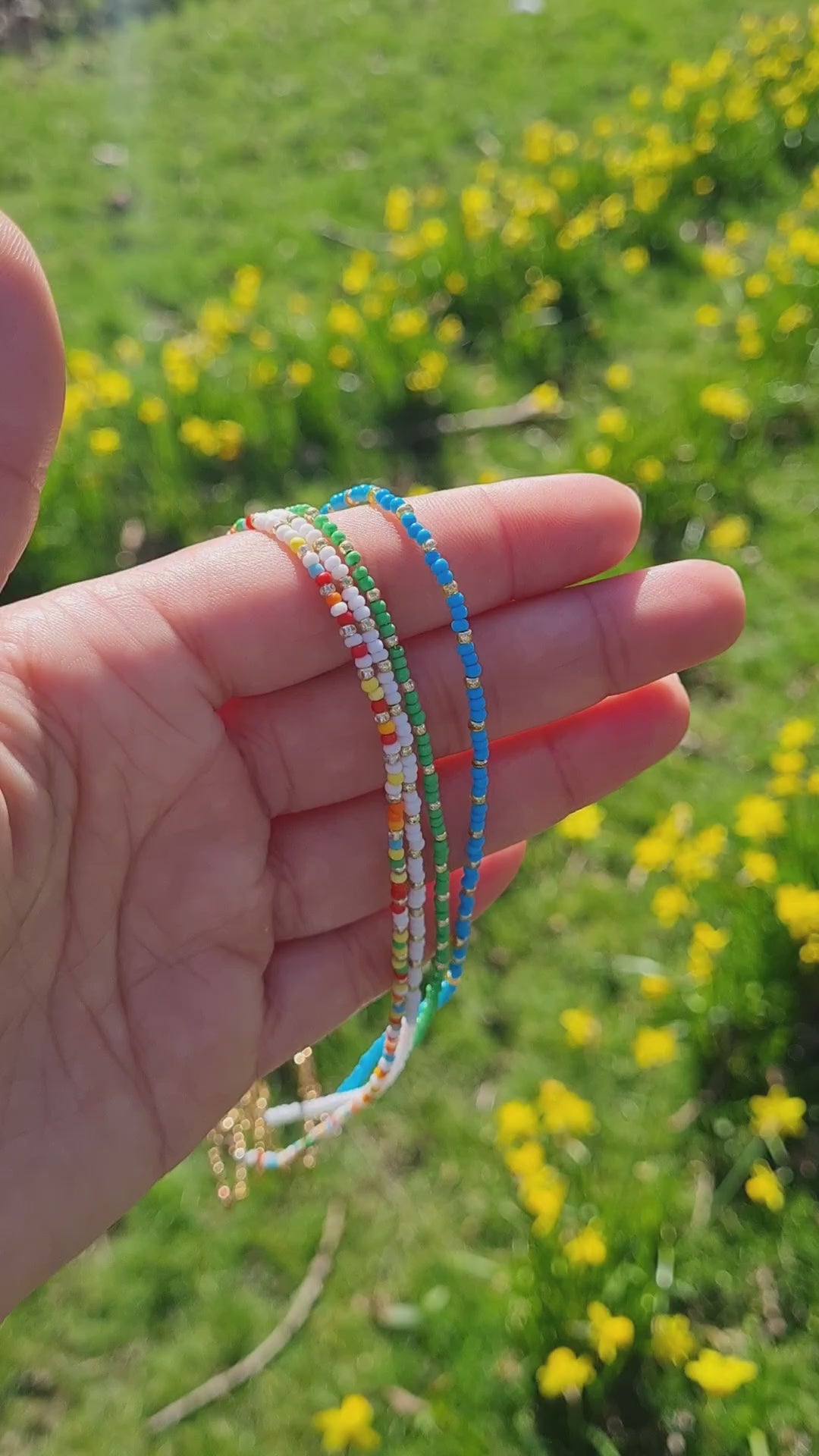 A mixture of handmade beaded necklaces with a gold plated chain s and a crystal miyuki beads being held up with a hand in the park.