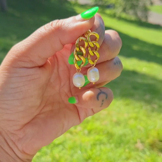 A hand holding a pair of 18K gold plated curb chain earrings with pearl drops, outdoor setting with grass and sunlight visible.