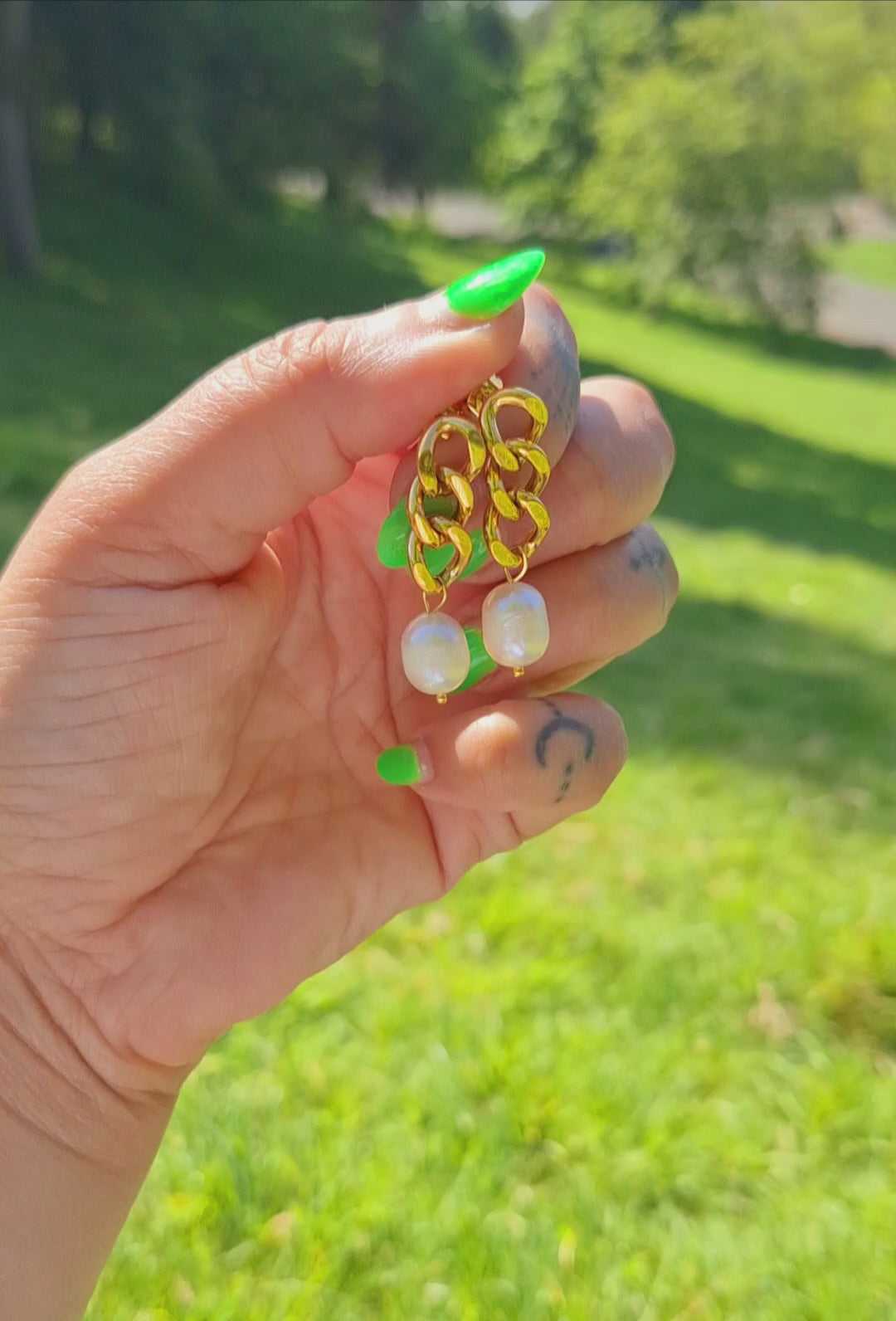 A hand holding a pair of 18K gold plated curb chain earrings with pearl drops, outdoor setting with grass and sunlight visible.